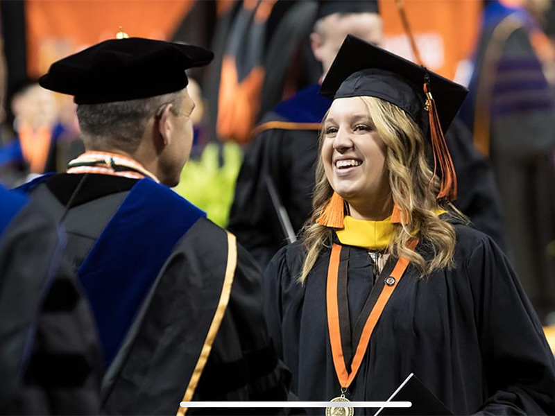A graduate student smiling widely and shaking hands with a professor at commencement