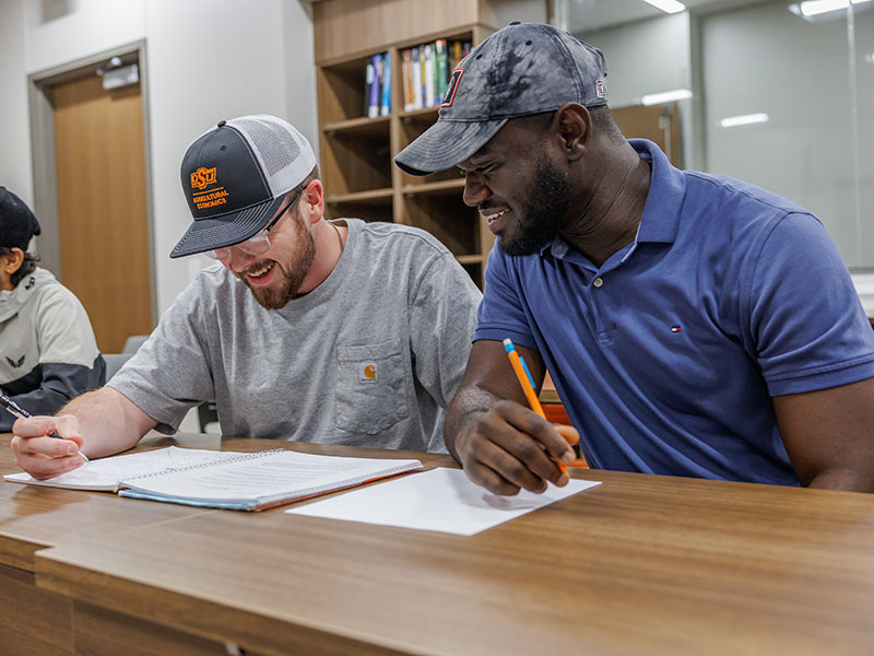 Two students sitting at their desks and looking at a notebook on the table.