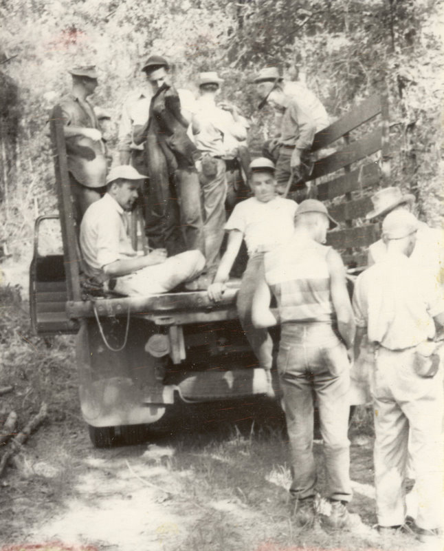 Students are standing and sitting in the back of a truck while others are standing around the back of the truck bed.