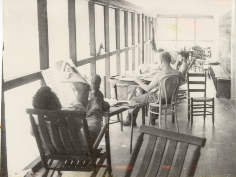 People are sitting on chairs on a porch with some reading a paper and others lounging.