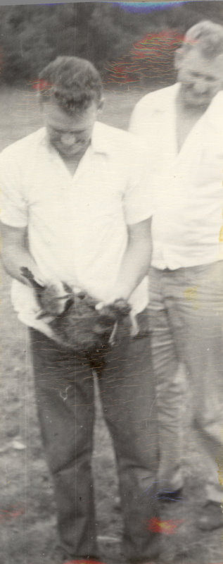 One man is holding a racoon while another man behind him looks down at the racoon.