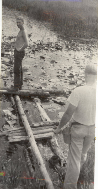 Two people are standing on logs over a creek.