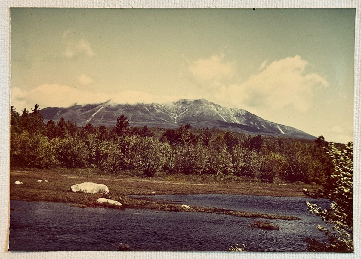 A blue river in the front, dark green trees in the middle and a mountain in the background.