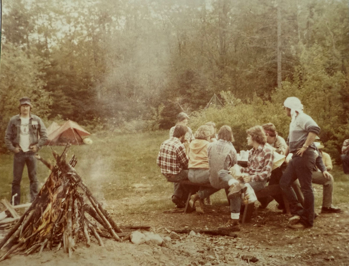 A group of people on the right sitting by a picnic table and a large fire on the left in the woods.