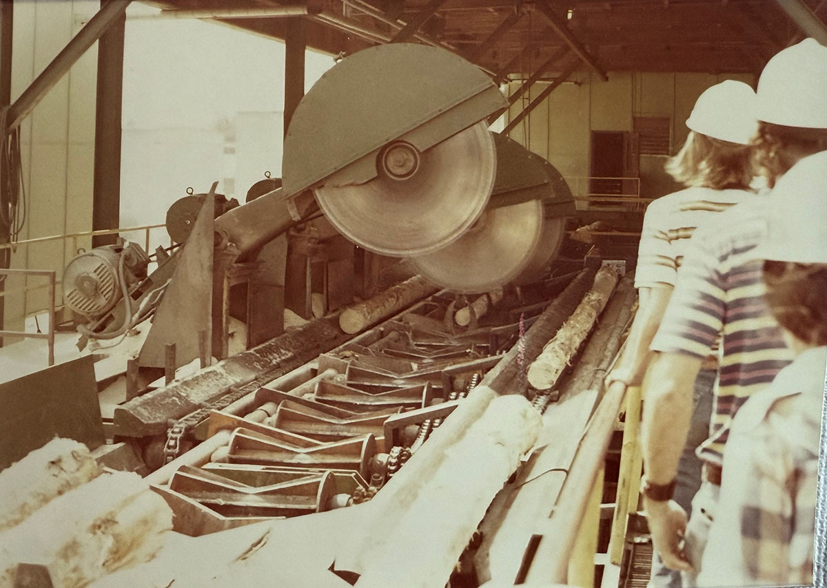 Large wooden log saws cutting small logs with students watching on the right.
