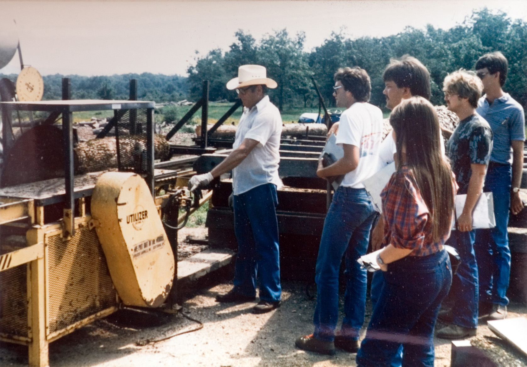 Five students watch an instructor operate yellow forestry equipment.