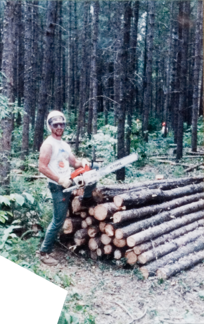 A student in a hardhat and a white shirt using a chainsaw to cut a stack of logs.