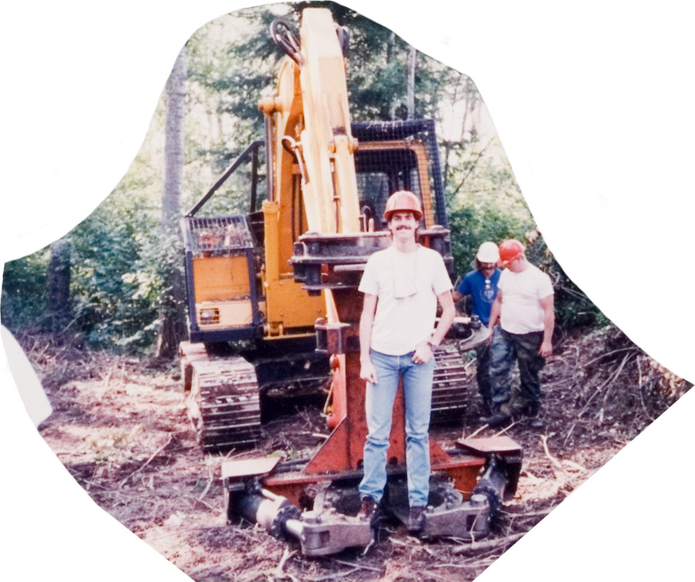 A student in a white shirt and red hardhat posing in front of yellow forestry equipment.