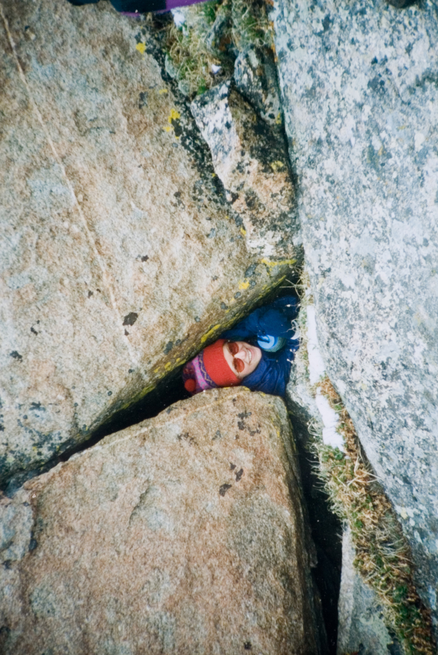 A student in an orange beanie smiling for the camera while poking their head out of a set of giant boulders.
