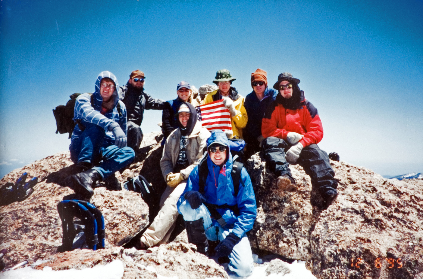 A group of eight students stitting on top of a boulder, while one holds an American flag for a photo.