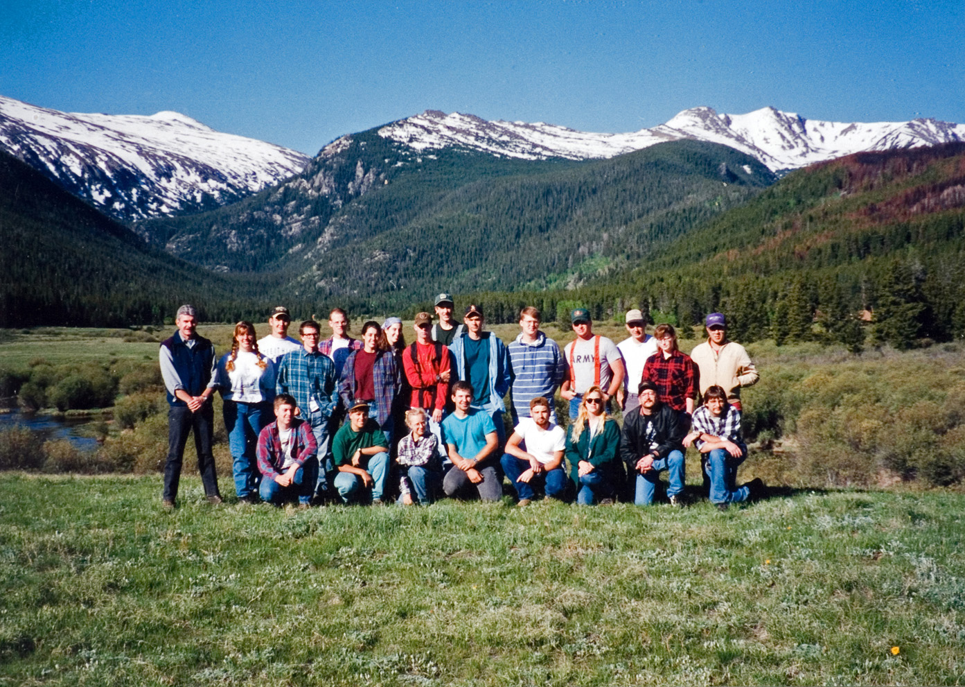 23 individuals from the forestry camp stand in a field of green grass with a mountain range behind them as they pose for a group photo.