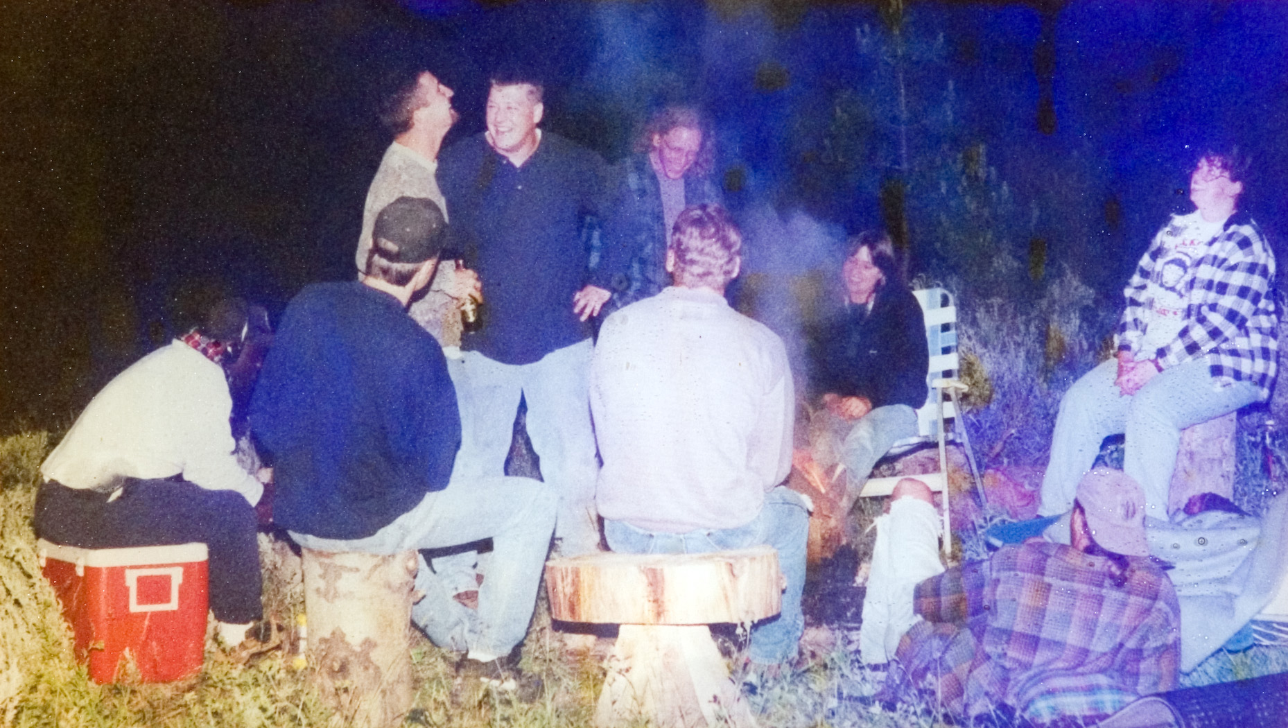 Nine students gathered outdoors around a campfire as some sit and some stand to convers with one another.