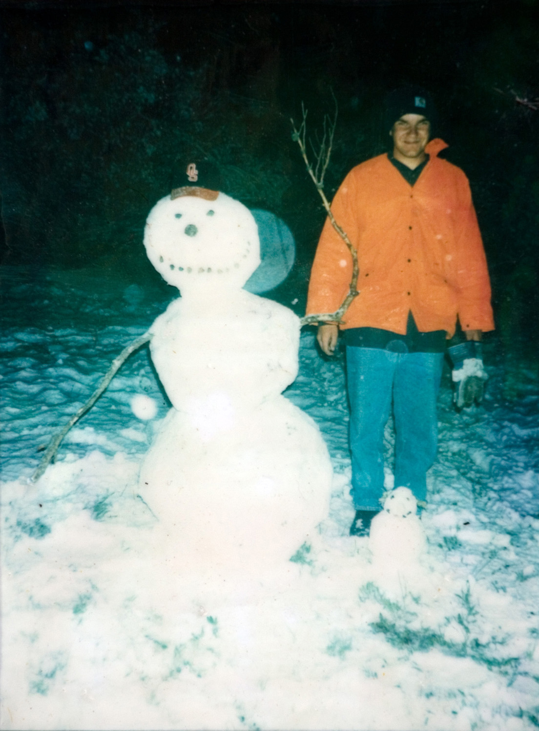 A student in an orange jacket standing next to a white snowman outside at night.