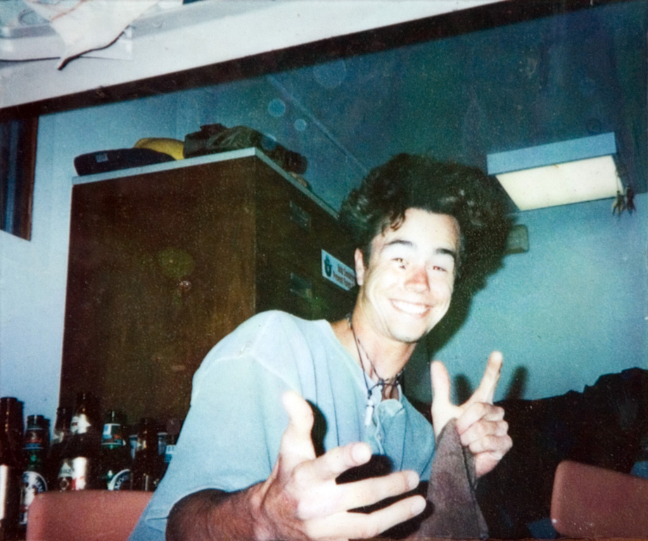 A student sitting indoors in a grey T-shirt smiles for his photo to be taken.