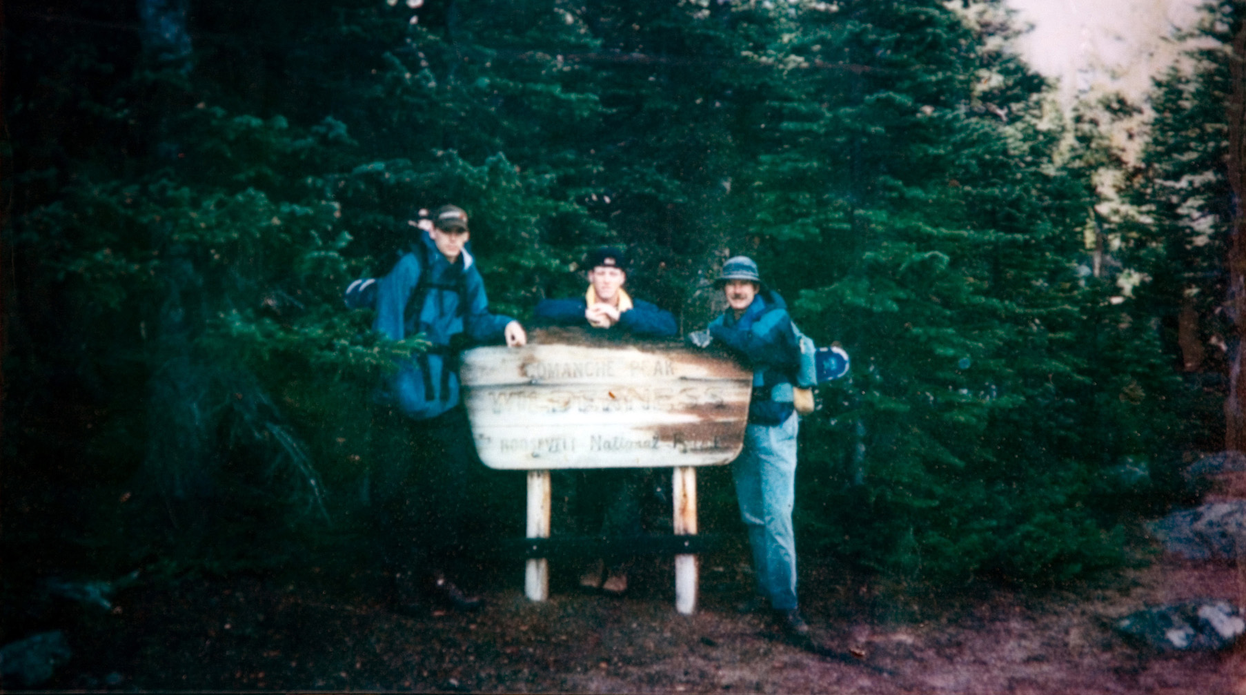 Three students standing behind a washed-out wooden sign as they pose in front of a forest of green trees for their photo to be taken.