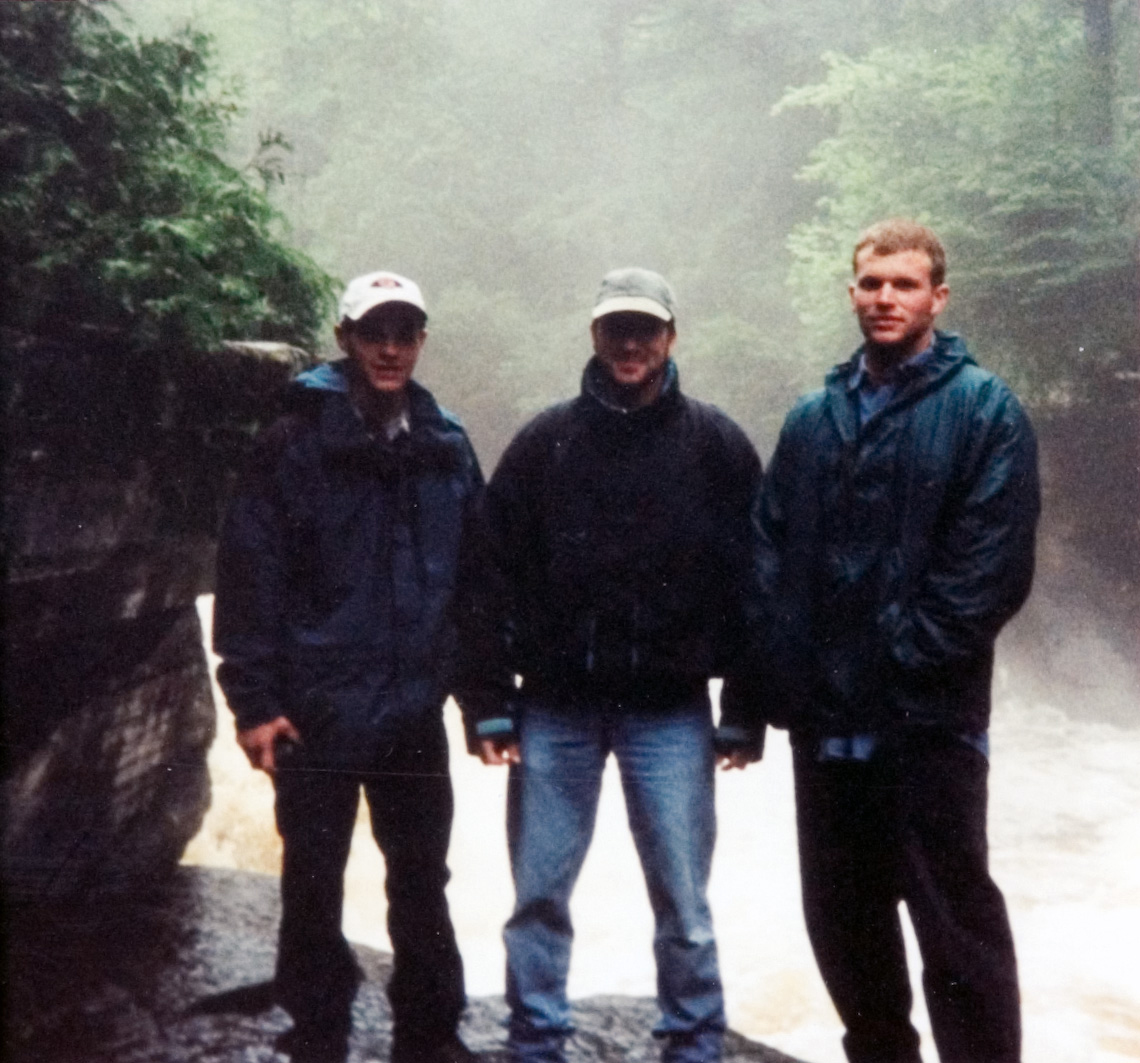 Three students standing in front of a misty stream of water as they pose for their photo to be taken.