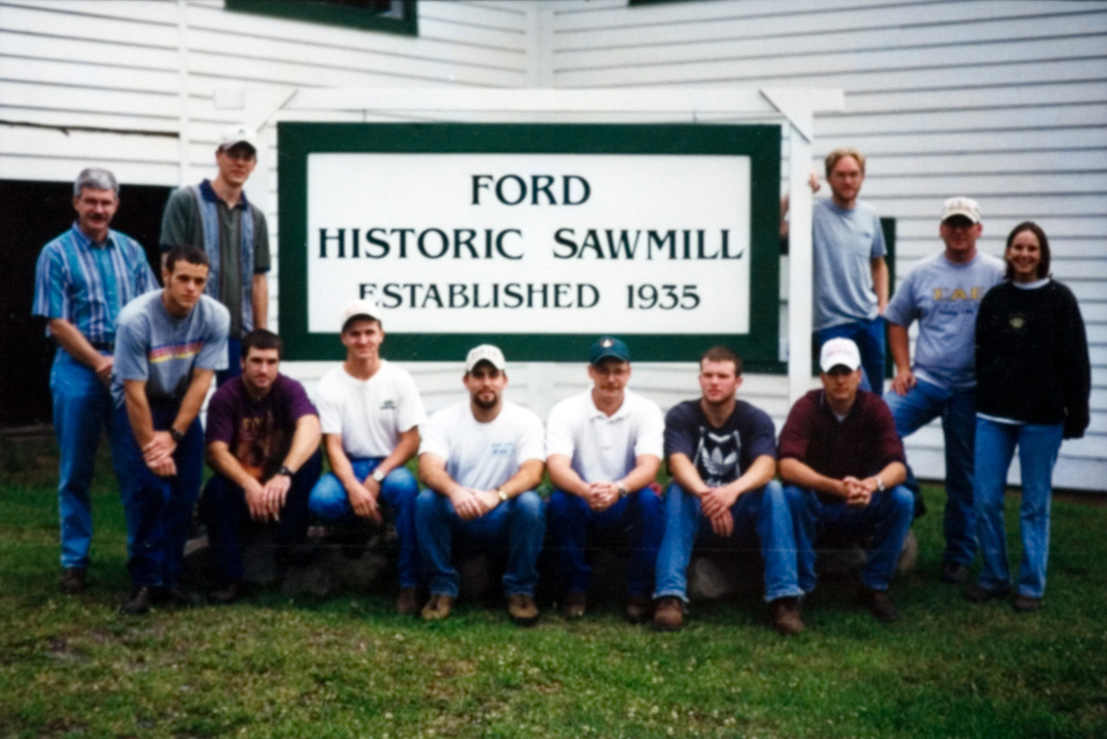 A group of students apart of the forestry camp standing in front of a sign that says "Ford Historic Sawmill Established 1935".