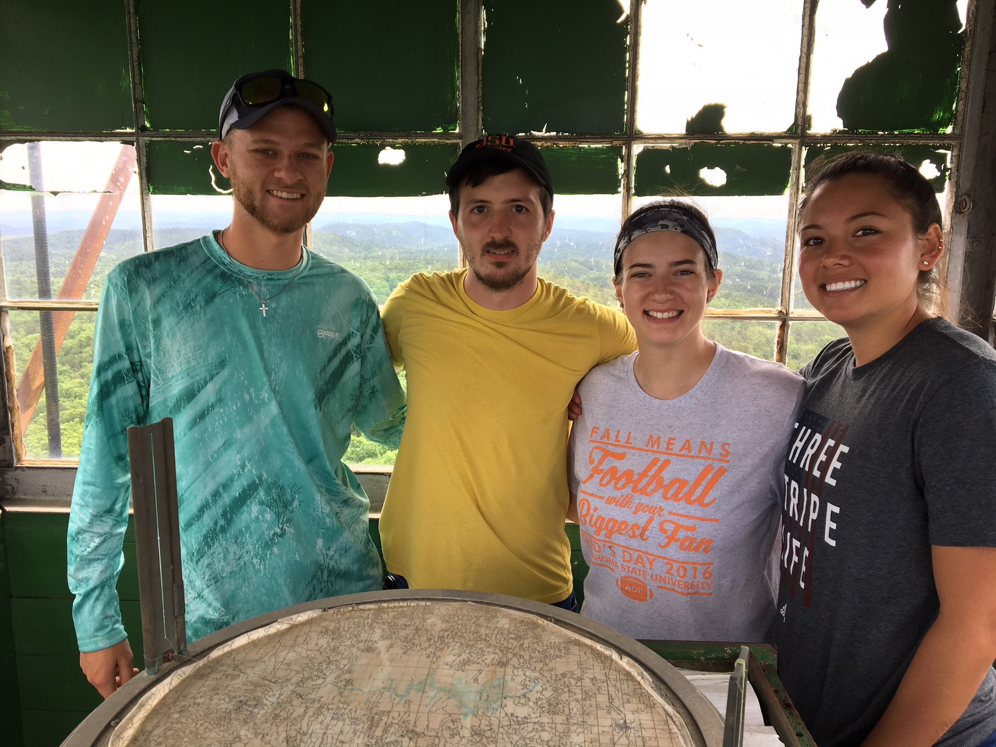 A group of four people standing at a round table, in the forest, smiling at the camera.