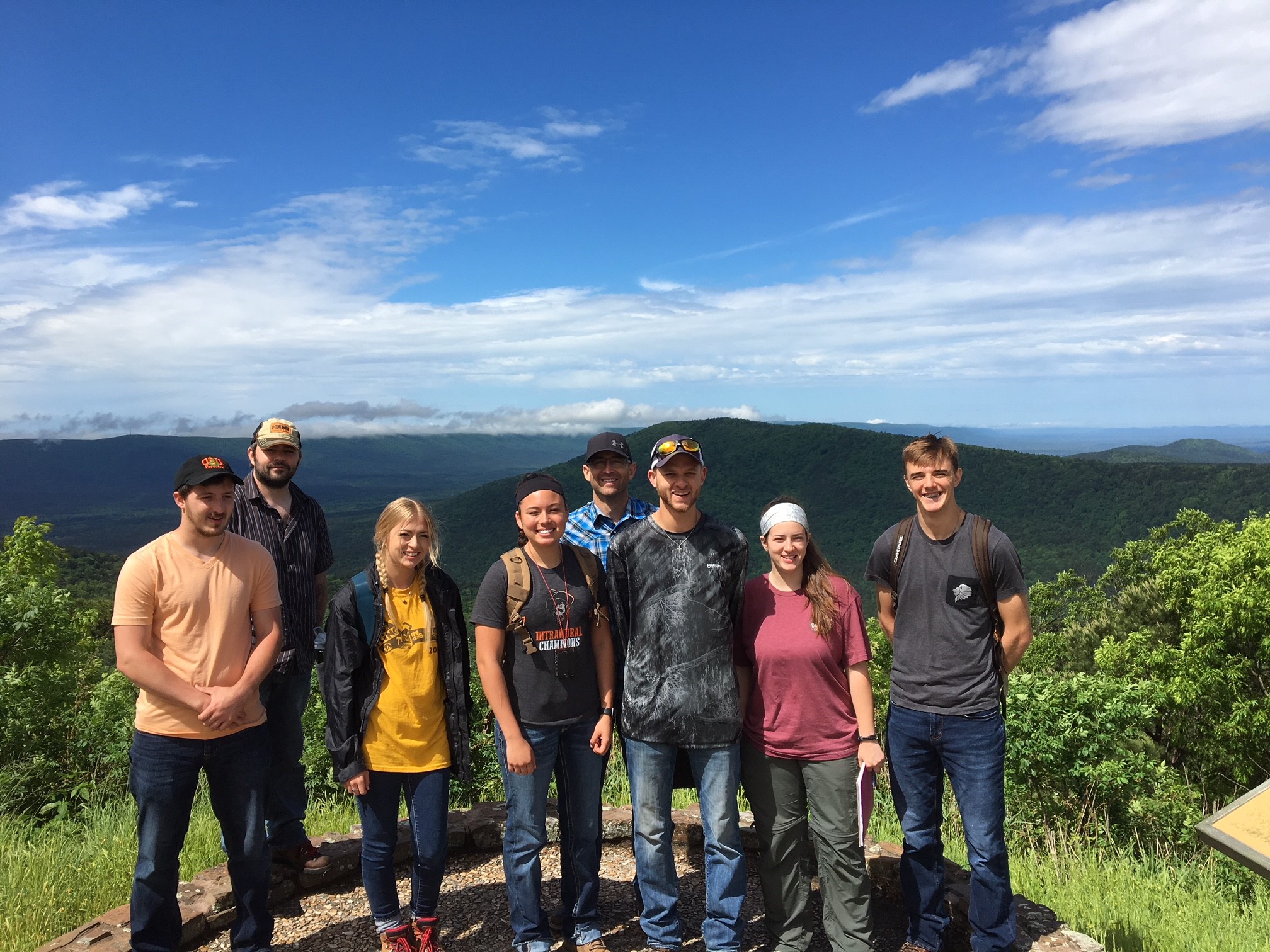 A group of eight individuals standing at the top of a mountain overlooking a forest.