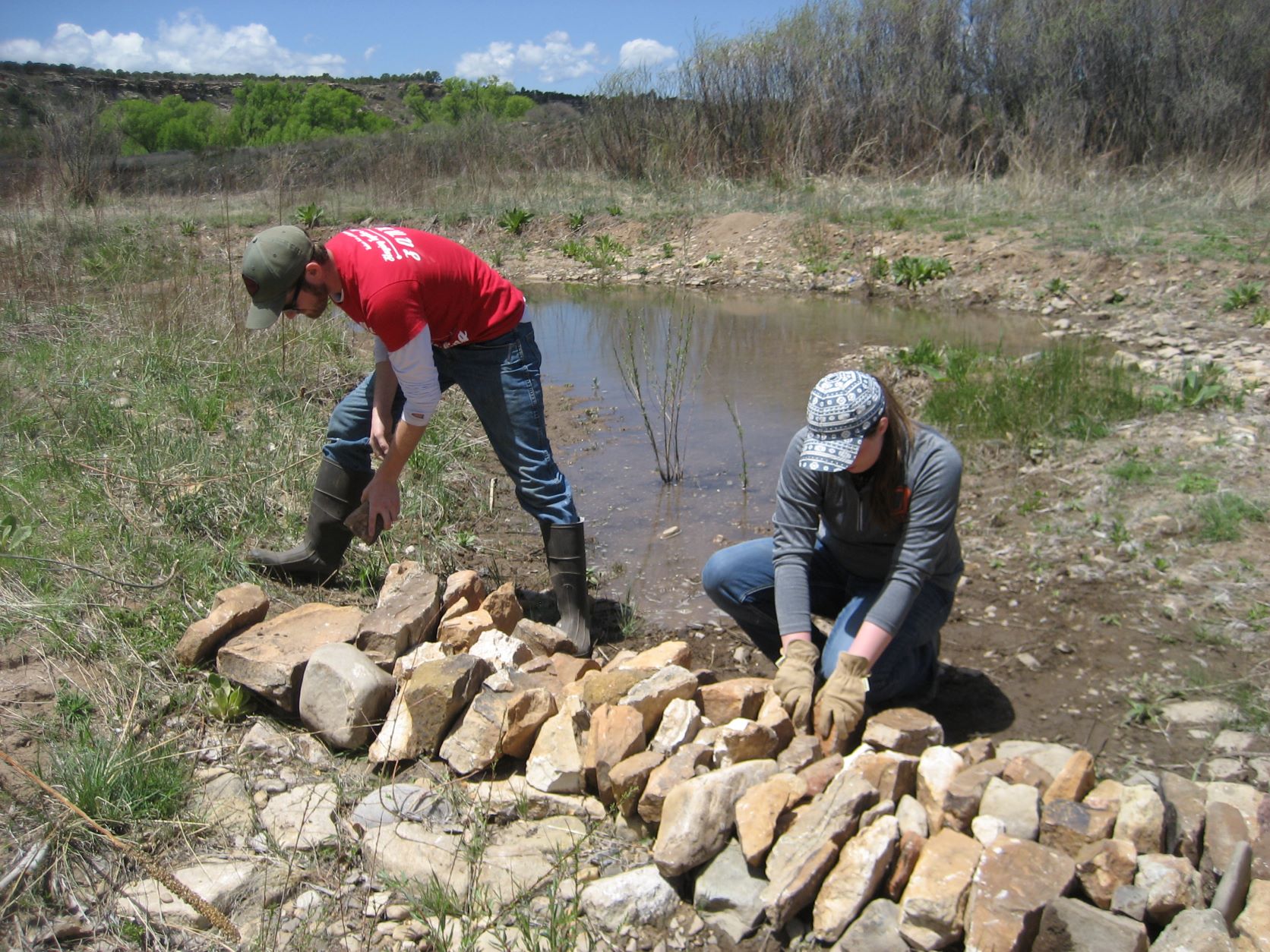 Two individuals arranging rocks beside a small pond in a valley.