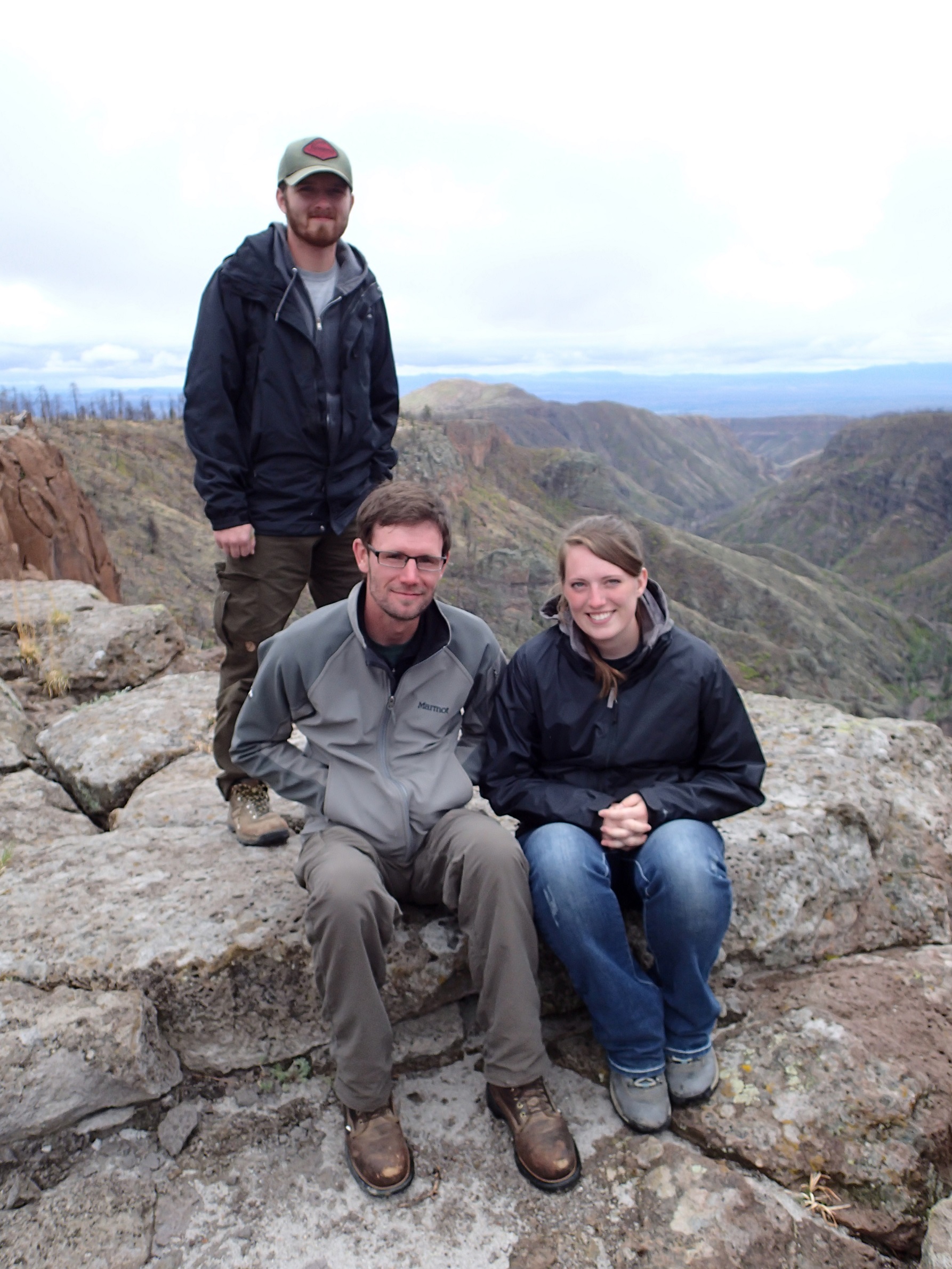 Three people at the top of a mountain overlooking the rest of the mountain range.