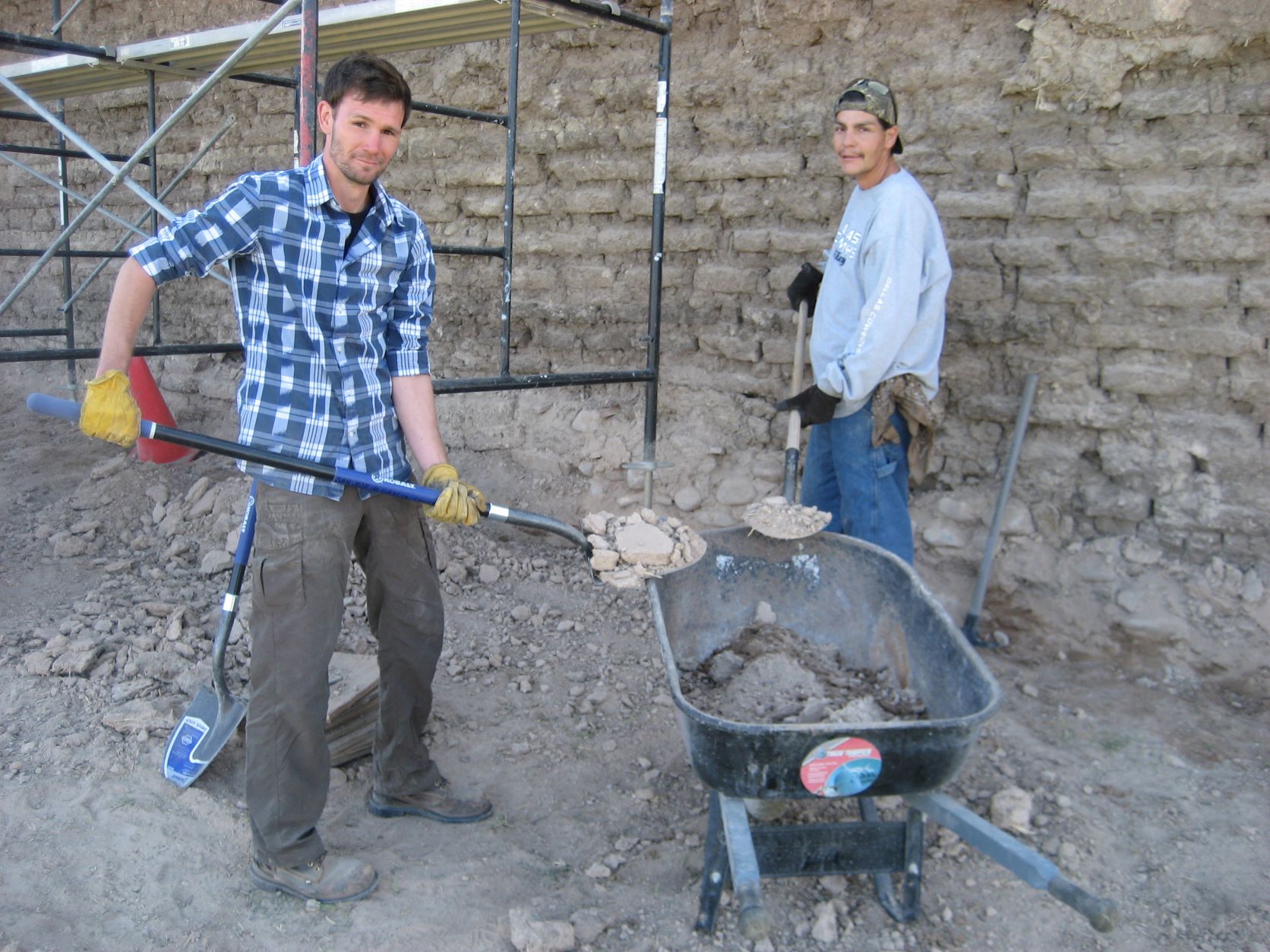 Two guys shoveling dirt and rocks into a wheelbarrow.