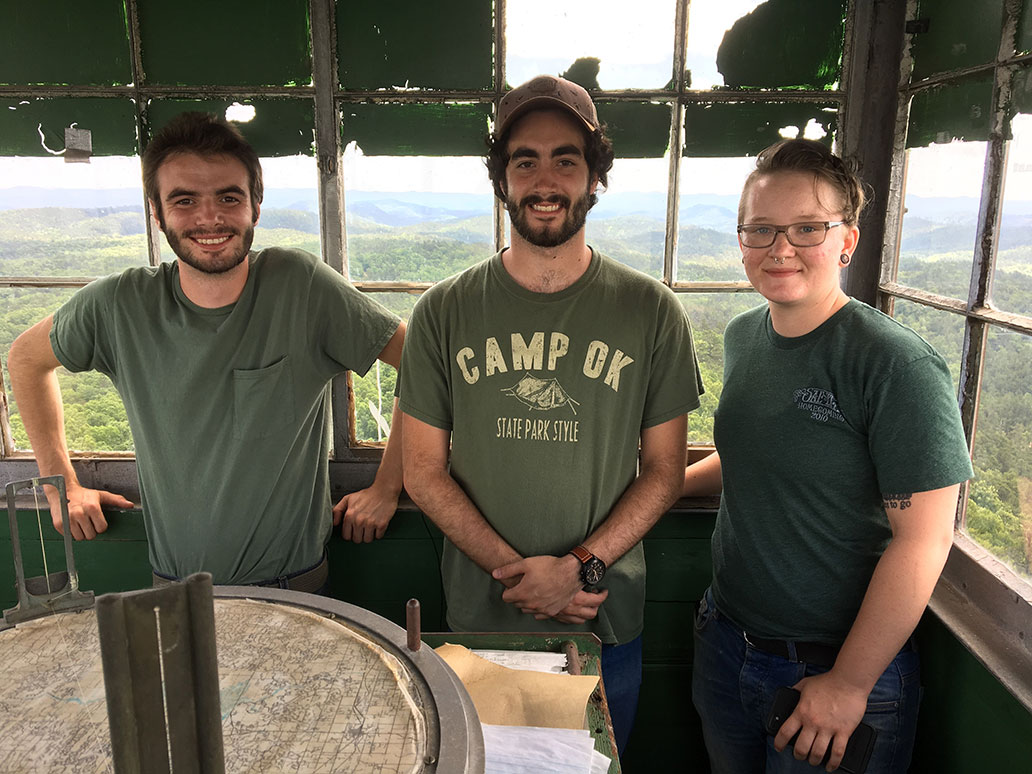 A group of three individuals standing at a round table in stand above the forest.