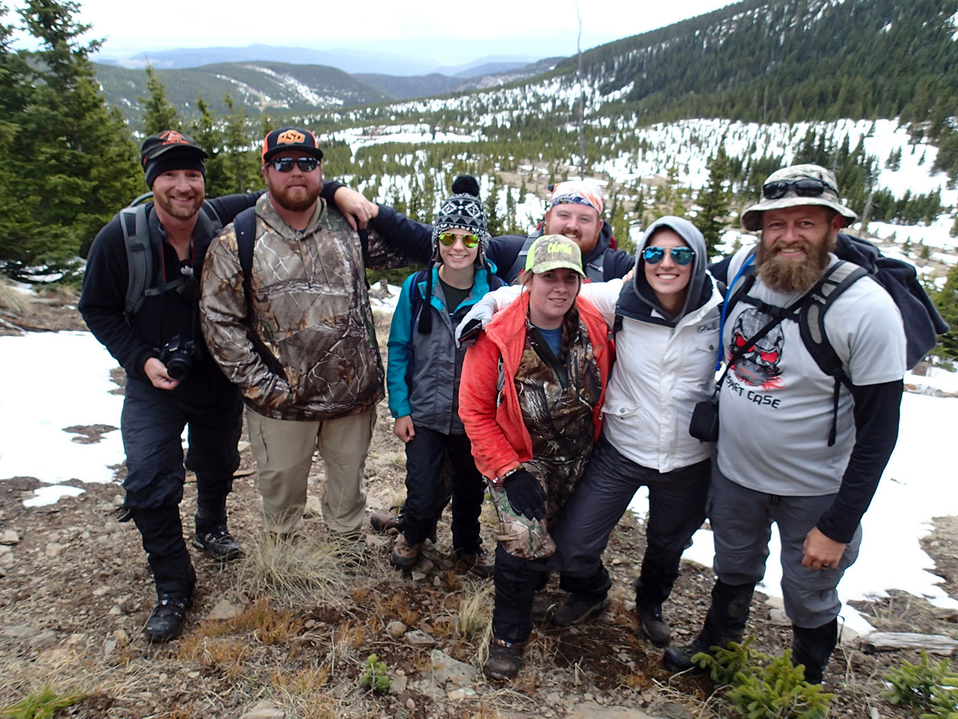 Seven individuals smiling at the camera while standing in the snow in a forest.