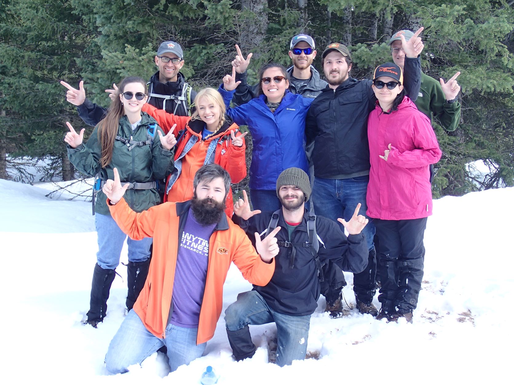 A group of 10 people in a snow-covered woods throwing the Oklahoma State "pistols firing" hand signals.