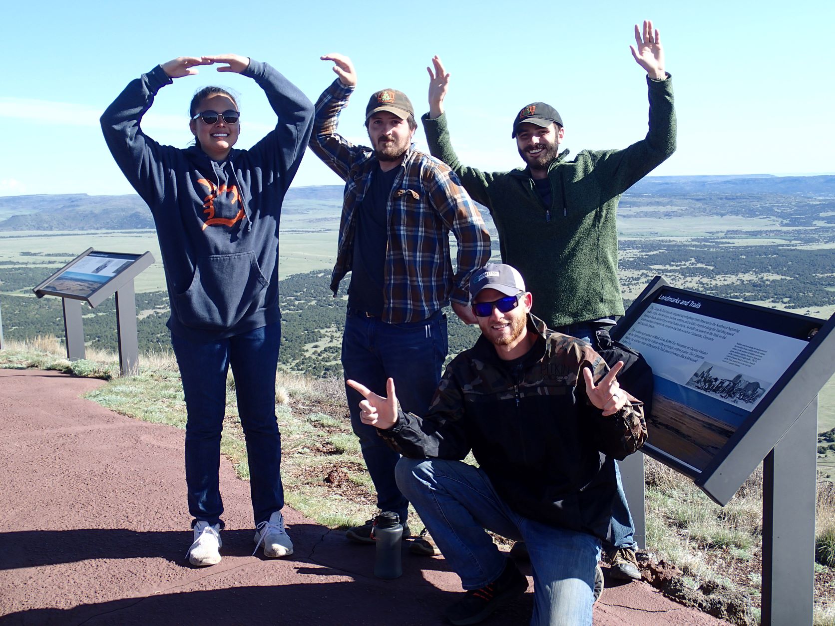 Four students creating the Oklahoma State "O.S.U" and "pistols firing" symbols with their arms at the top of a mountain overlook.