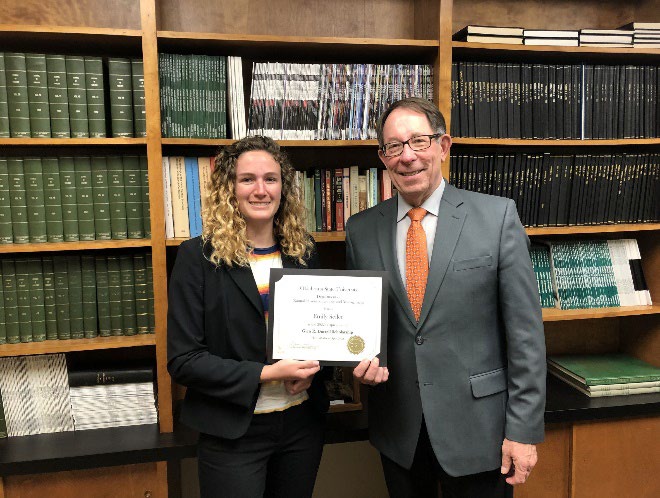 Emily Swiler is accepting her award from Jim Ansley in front of a book shelf.