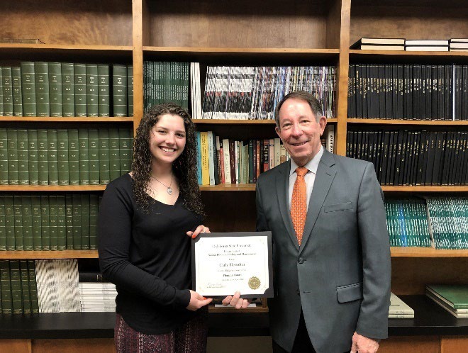 Carly Herndon is accepting her award from Jim Ansley in front of a book shelf.