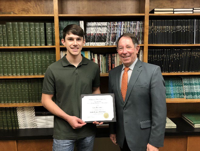 Coy Brewster is accepting his award from Jim Ansley in front of a book shelf.