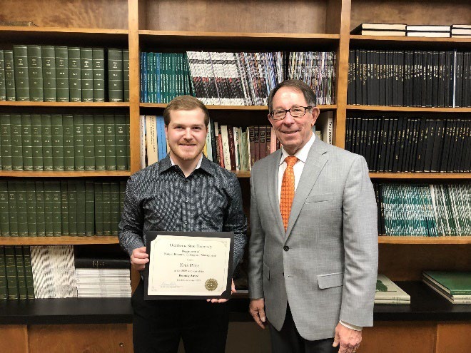 Evan Price is accepting his award from Jim Ansley in front of a book shelf