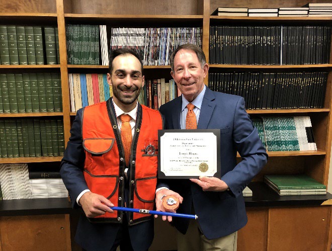 Loseph Hogan is accepting his award with a ornage west on from Jim Ansley in front of a book shelf