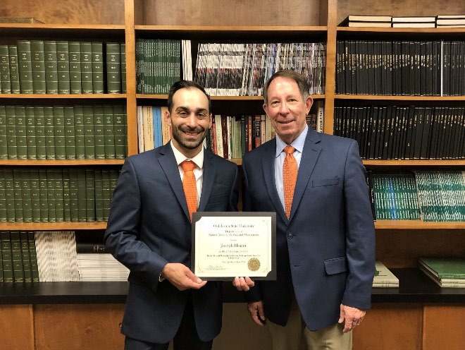 Loseph Hogan is accepting his award from Jim Ansley in front of a book shelf