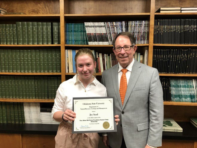 Joy Sand is accepting her award from Jim Ansley in front of a book shelf.