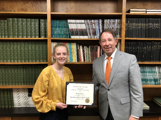 Kendall Hays is accepting her award from Jim Ansley in front of a book shelf.