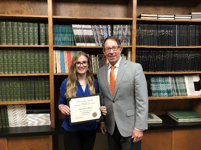 Mackenzie Hiberd is accepting her award from Jim Ansley in front of a book shelf.