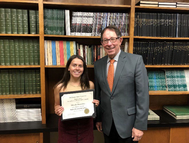 Mikayla Humphrey is accepting her award from Jim Ansley in front of a book shelf.