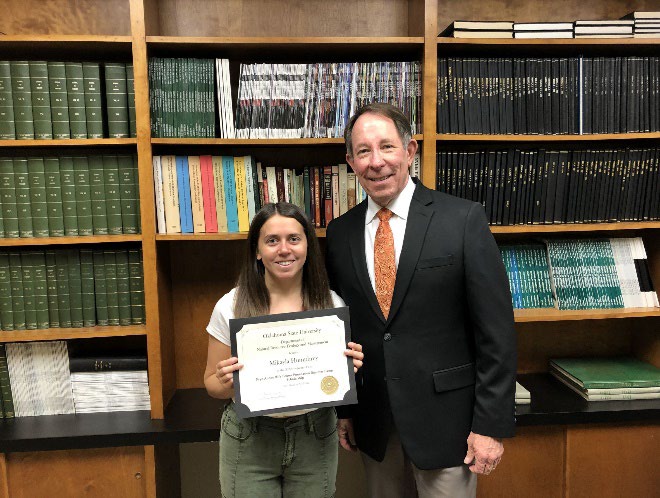 Mikayla Humphrey is accepting her award from Jim Ansley in front of a book shelf.