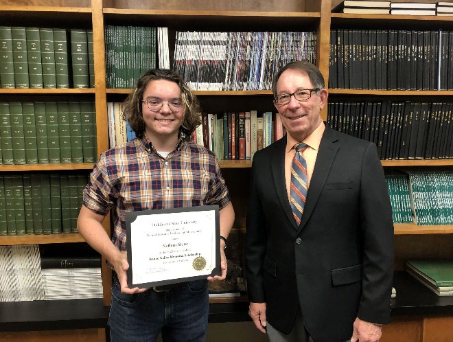 Nathan Stene is accepting his award from Jim Ansley in front of a book shelf