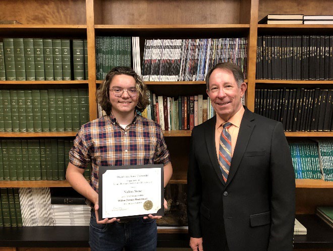Nathan Stene is accepting his award from Jim Ansley in front of a book shelf.
