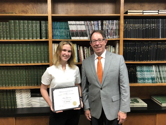 Olivia Horton is accepting her award from Jim Ansley in front of a book shelf.