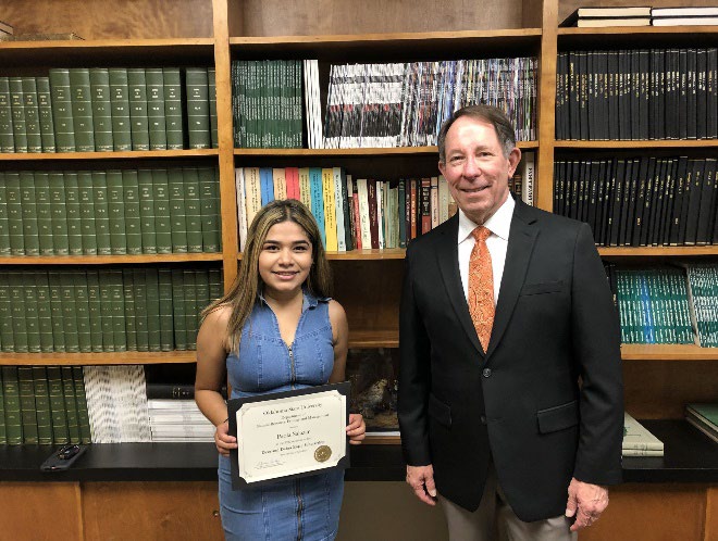 Paola Salazar is accepting her award from Jim Ansley in front of a book shelf.