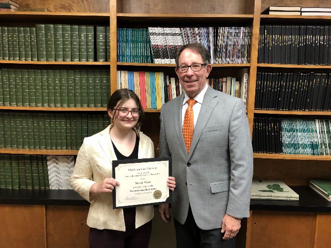 Maddi Watts is accepting her award from Jim Ansley in front of a book shelf.