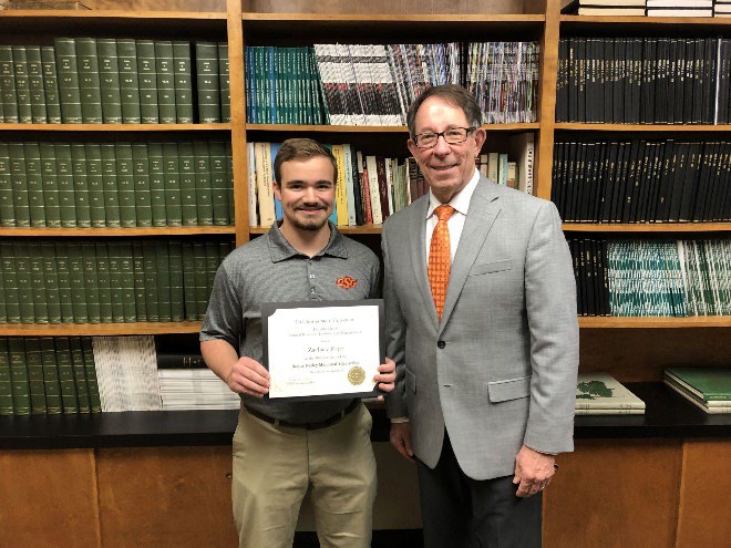 Zachary Pope is accepting his award from Jim Ansley in front of a book shelf