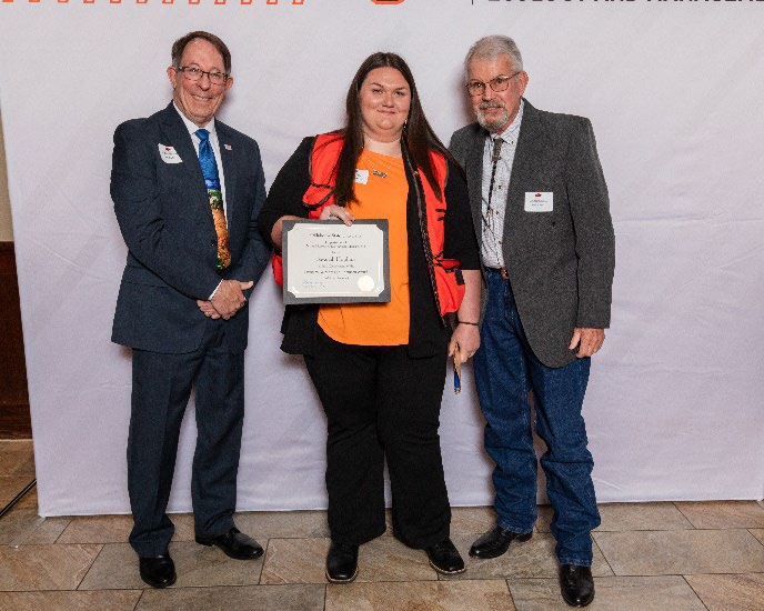 Savanah has a orange vest on and holding her award next to Tom and Jim.